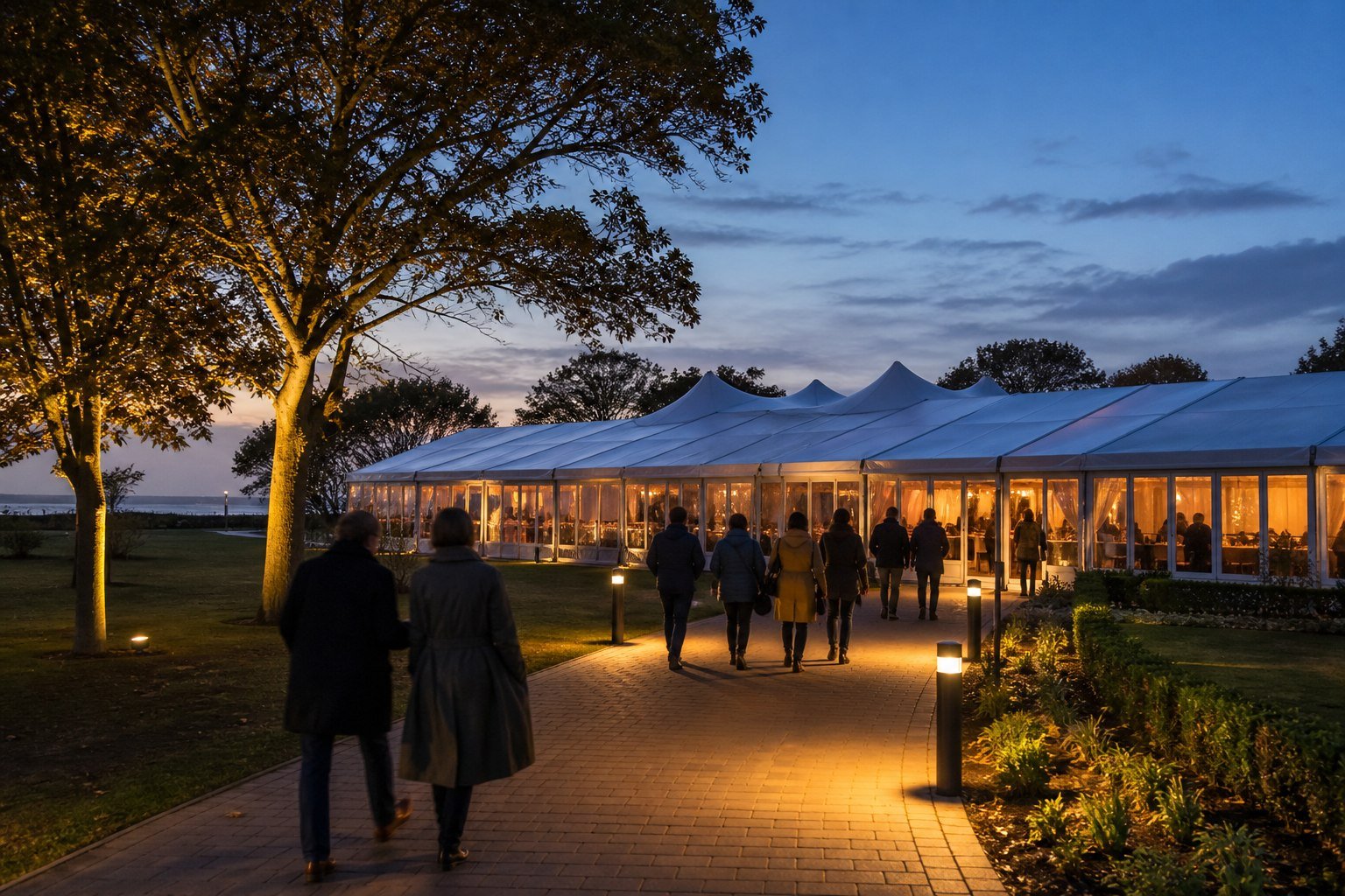 Visitors arriving at a warmly lit event marquee in a British park at dusk